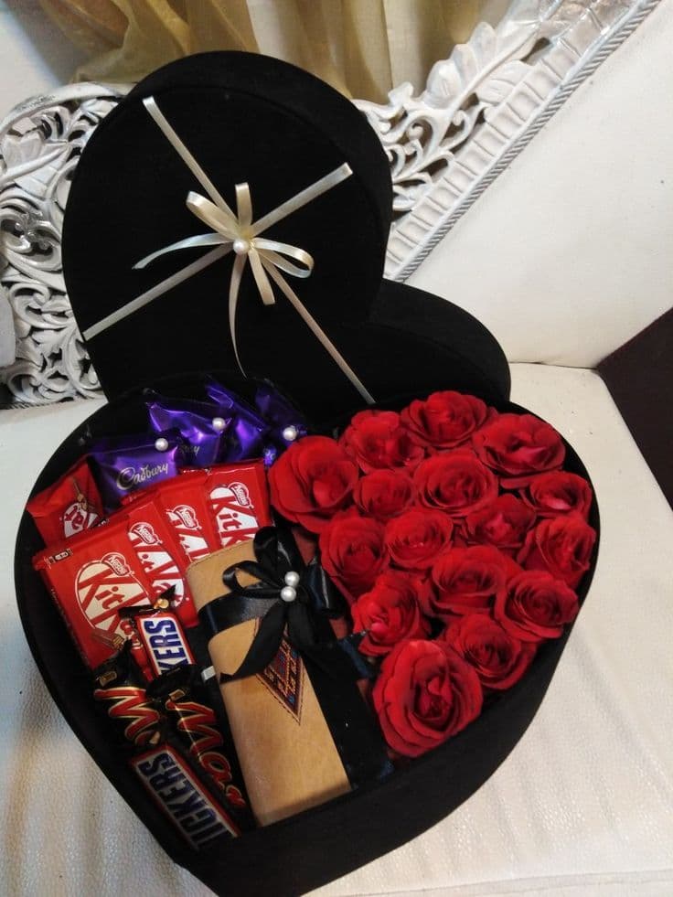 Black heart-shaped box filled with red roses, assorted chocolates, and a small wrapped gift.