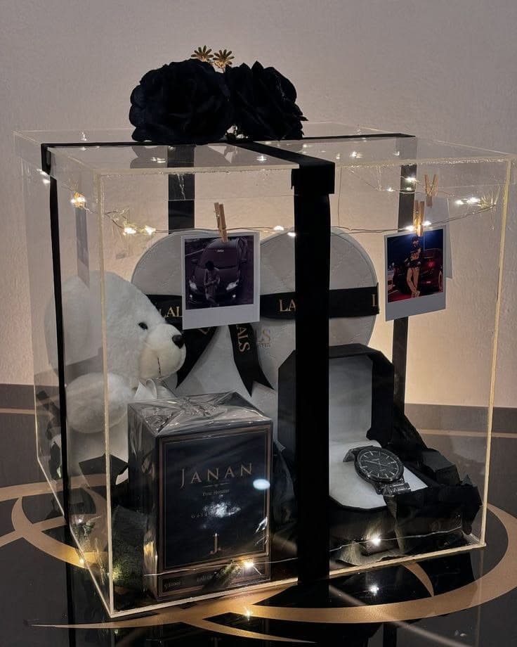 Clear gift box containing a white teddy bear, perfume, watch, and photos with fairy lights.